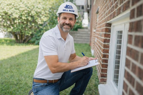 Sell Now Solutions cash buyer in a branded hard hat assessing a brick home with a clipboard